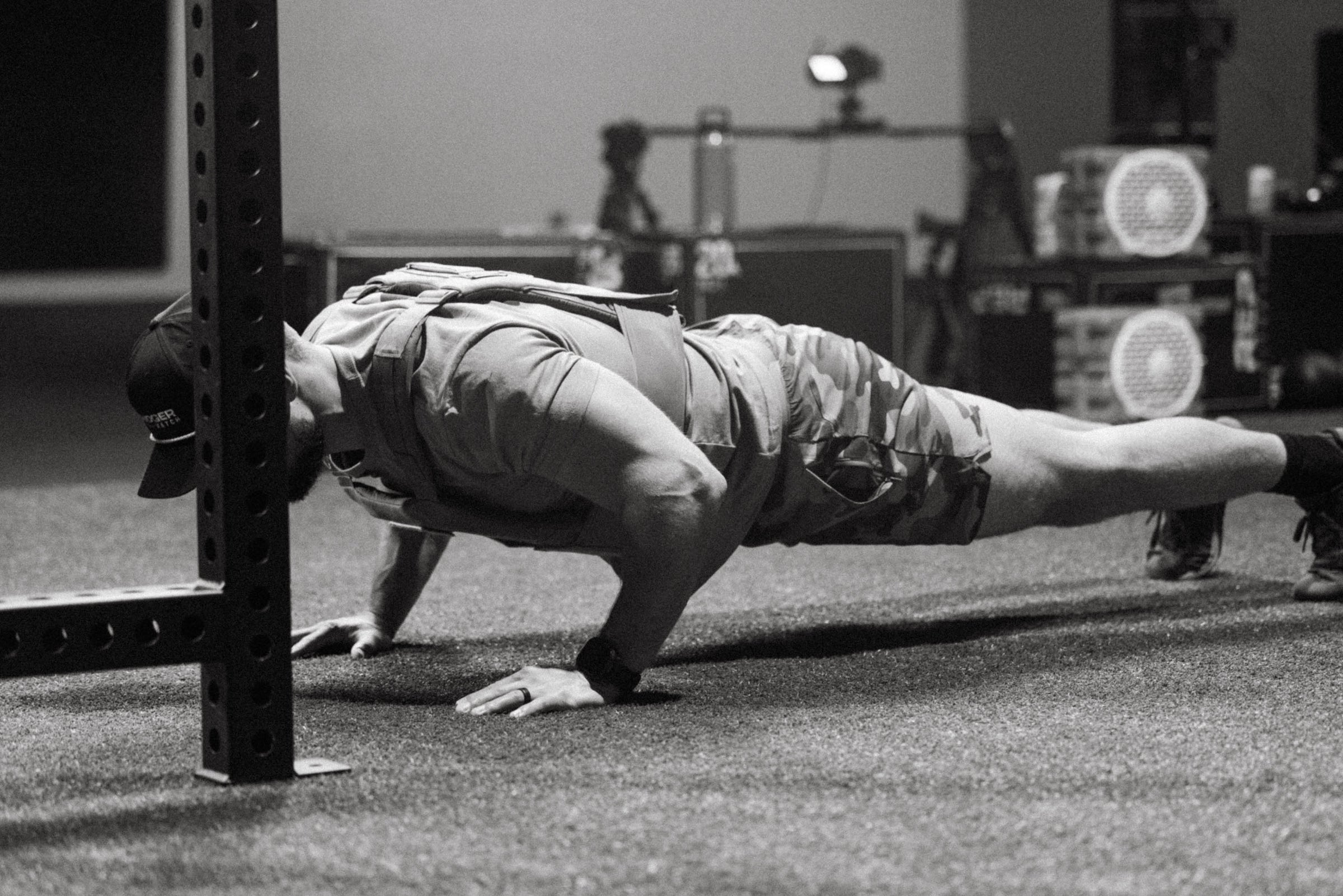 A man in a push-up position wearing a tactical vest and camouflage shorts in a gym setting, captured in a black and white photograph.