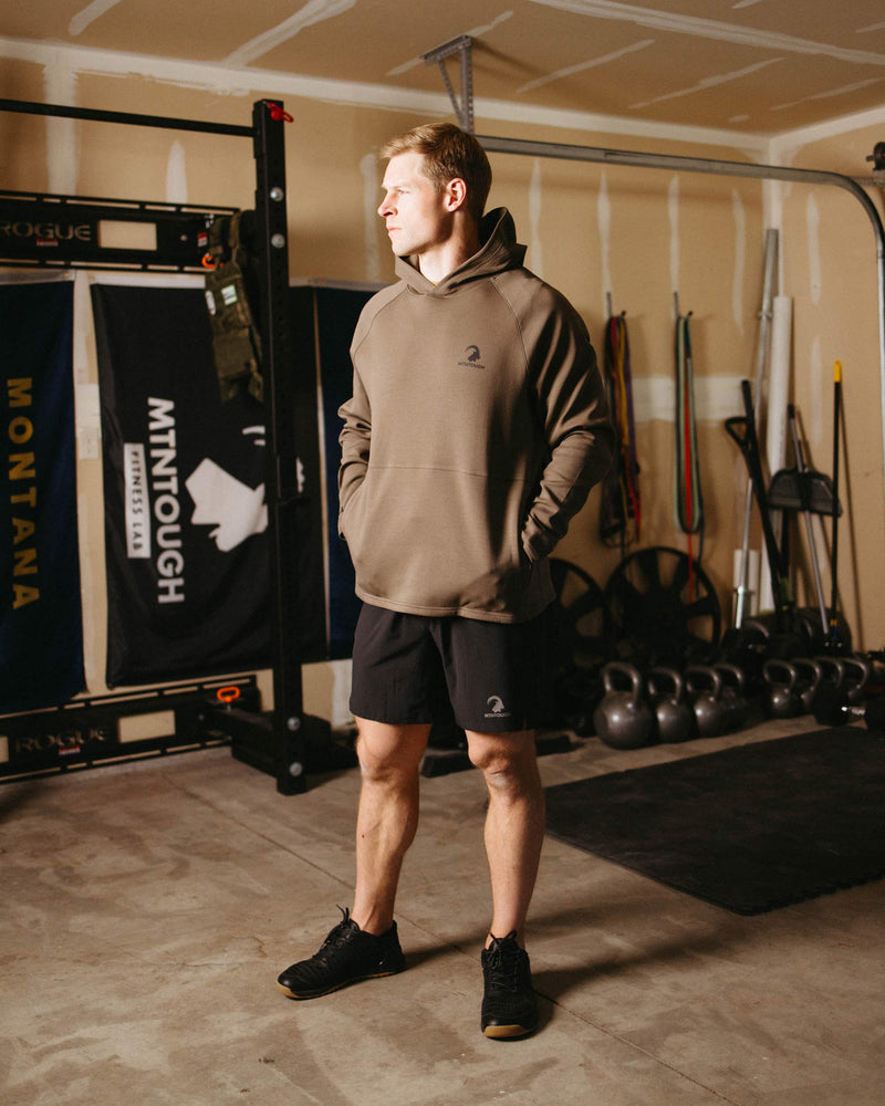 Man in a gym wearing a brown hoodie and black shorts, standing in front of gym equipment.