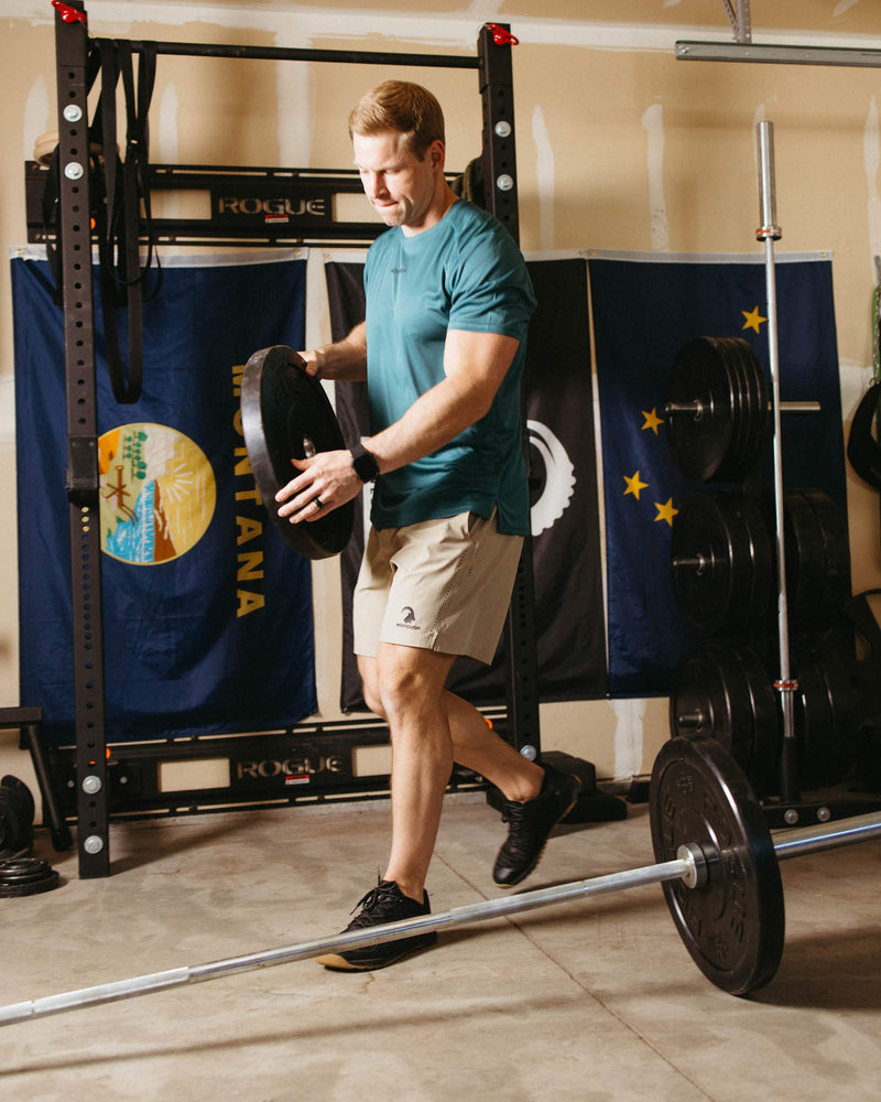 Man lifting a barbell in a home gym setting with equipment and flags in the background.