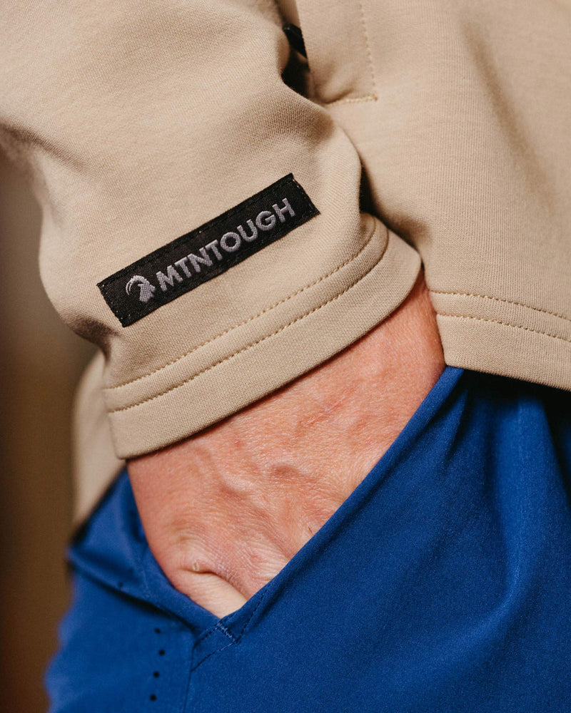 Close-up of a beige sleeve with a '2MTNTOUGH' label, worn by a person in a blue shirt.